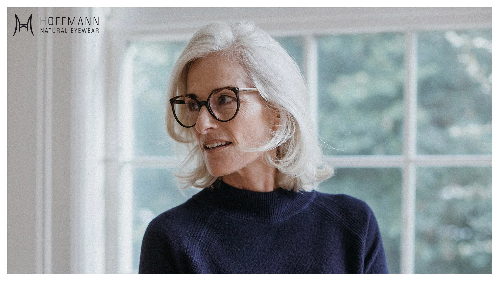 Nahaufnahme von einem älteren Female Model mit schulterlangen, weißen Haaren vor einem Fenster stehend, das eine braune Hoffmann Natural Eyewear Brille trägt.