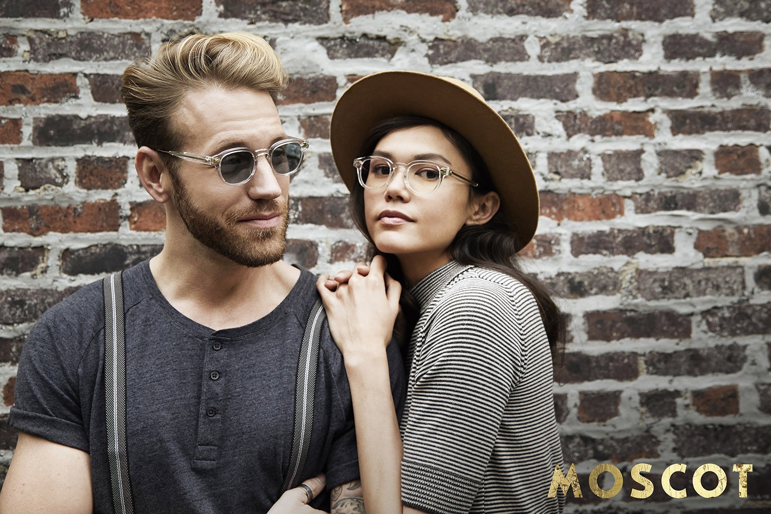 Ein weibliches Model rechts und ein männliches Model links tragen Moscot Eyewear stehen vor einer Bausteinwand.