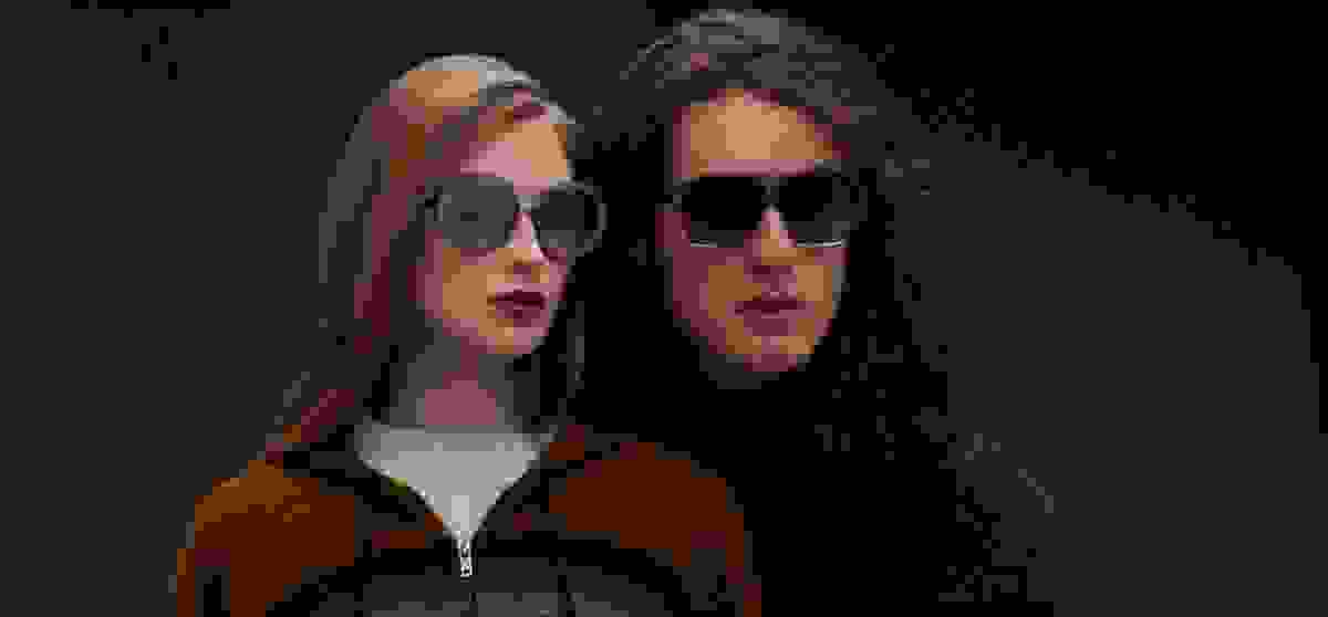 Nahaufnahme mit schwarzem Hintergrund von einer Frau mit rötlichen Haaren und einem Mann mit langen Haaren, die Blake Kuwahara Sonnenbrillen tragen und in die Ferne schauen.