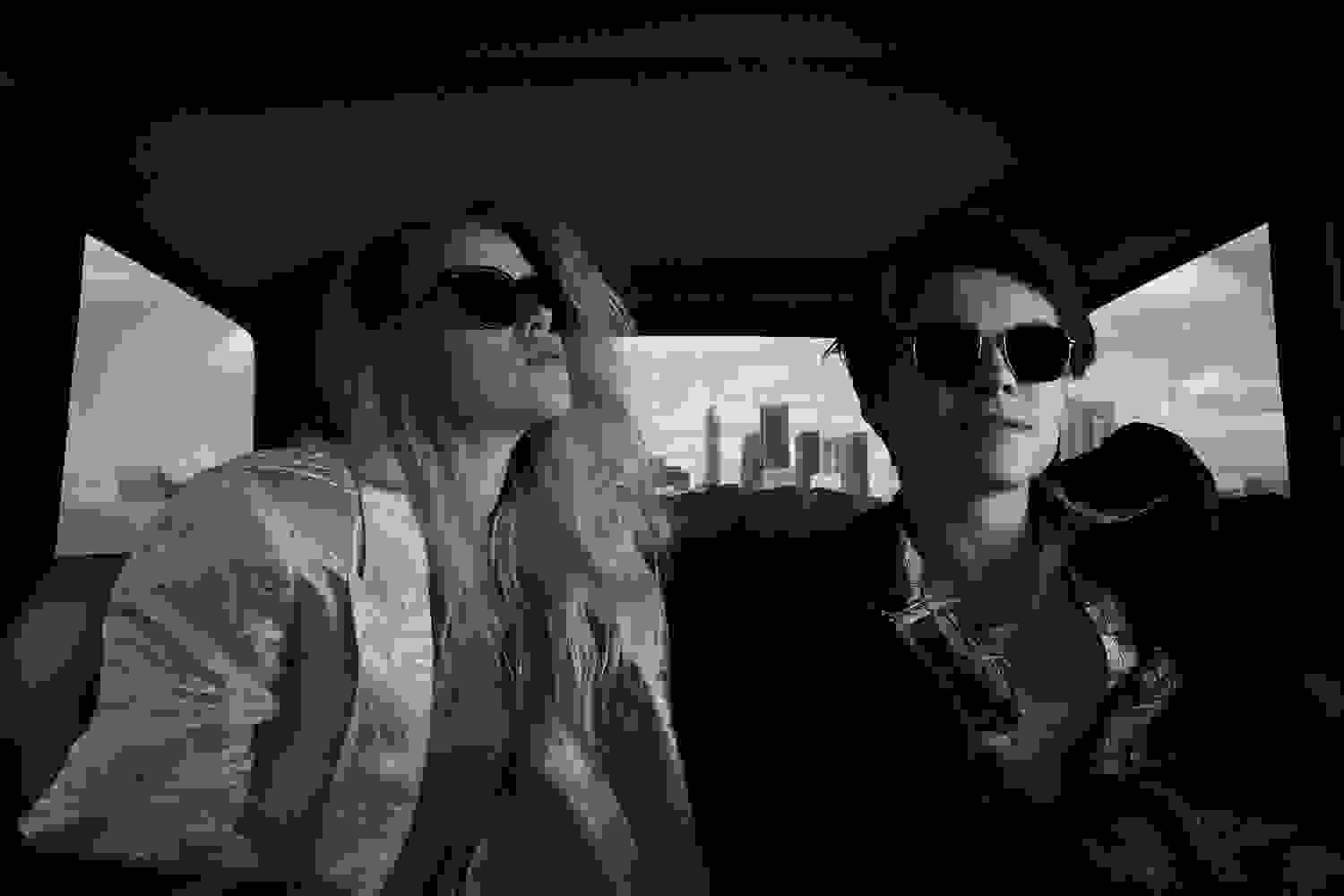 Schwarz-weiße Aufnahme von einem weiblichen und einem männlichen Model im Auto, die dunkle Sonnenbrillen tragen.