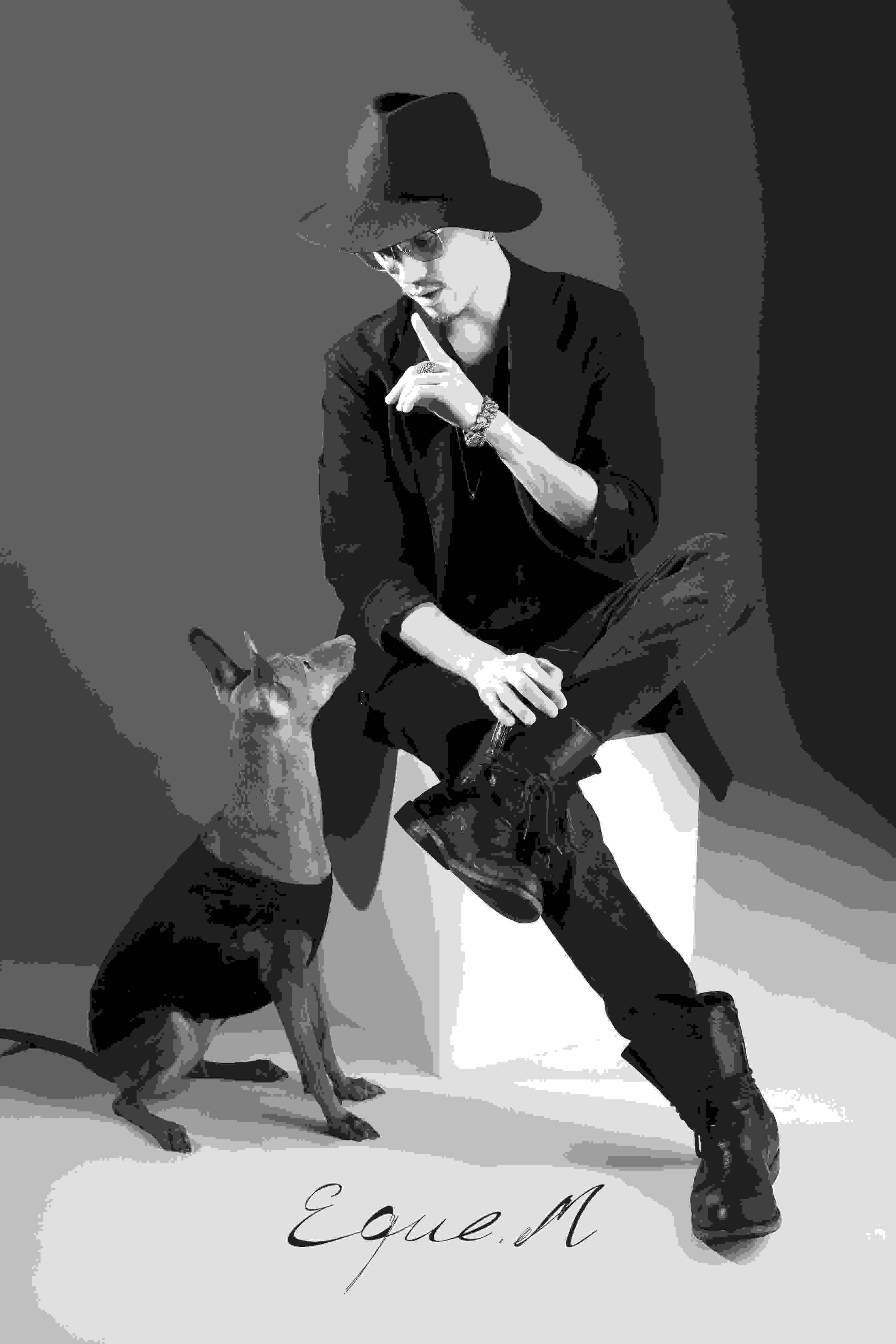 Schwarz-weiß Aufnahme von einem Mann mit schwarzer Kleidung und Hut, mit dem Blick zu einem auf dem Boden sitzenden Hund gerichtet.